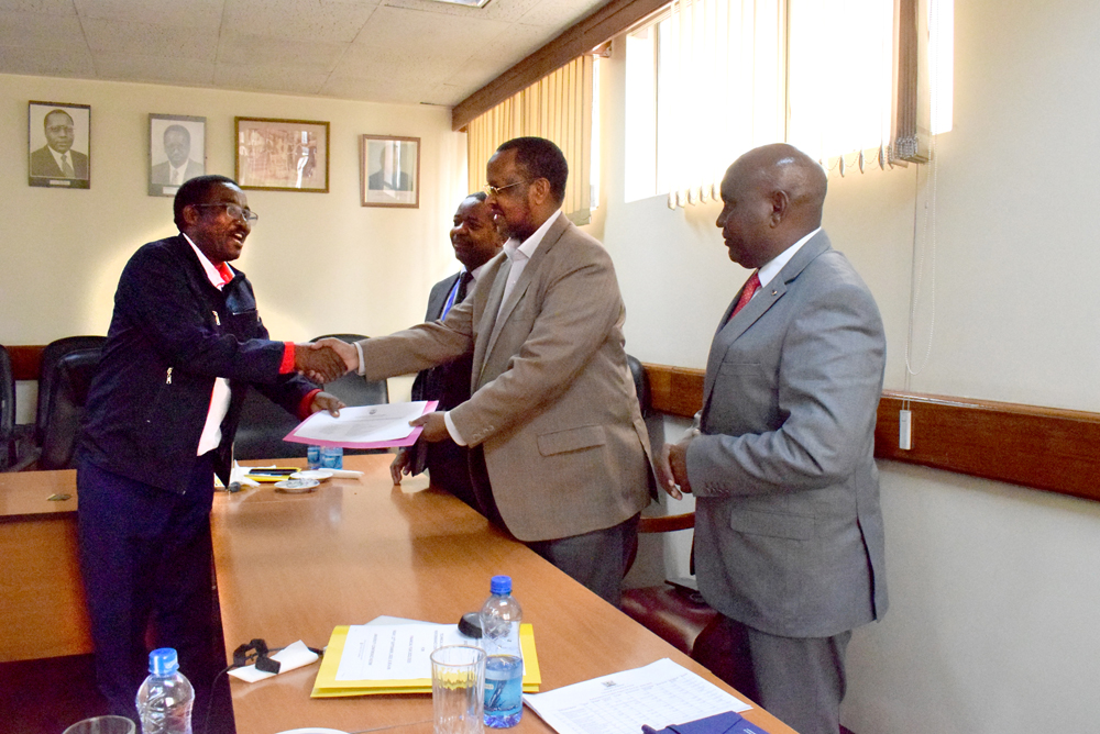 Mr Peter Kariuki from the Public Service Performance Management Unit hands the PC scoresheet to the Chairman of the University Council, Dr Idle Farah as a Council member, Hon. David Koech as the Vice-Chancellor, Prof Benedict Mutua looks on.